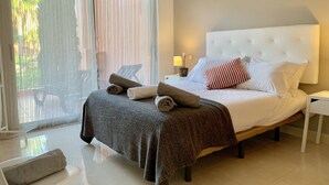 Une chambre à coucher avec un grand lit, une table de chevet, une lampe et une vue sur l’extérieur à travers une fenêtre ornée de rideaux légers.