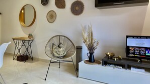 Un salon moderne avec un miroir rond, des œuvres d’art décoratives accrochées au mur, un fauteuil en osier, un canapé blanc, un téléviseur et une bibliothèque.