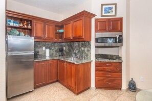 A compact kitchen with wooden cabinets, a stainless steel refrigerator, a microwave, and a granite countertop.