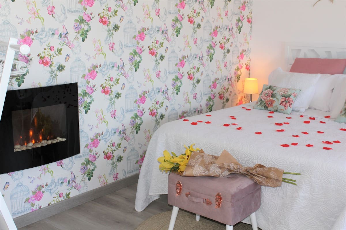 A bedroom with floral wallpaper, a bed with rose petals, a bedside table with a bouquet, and a fireplace.