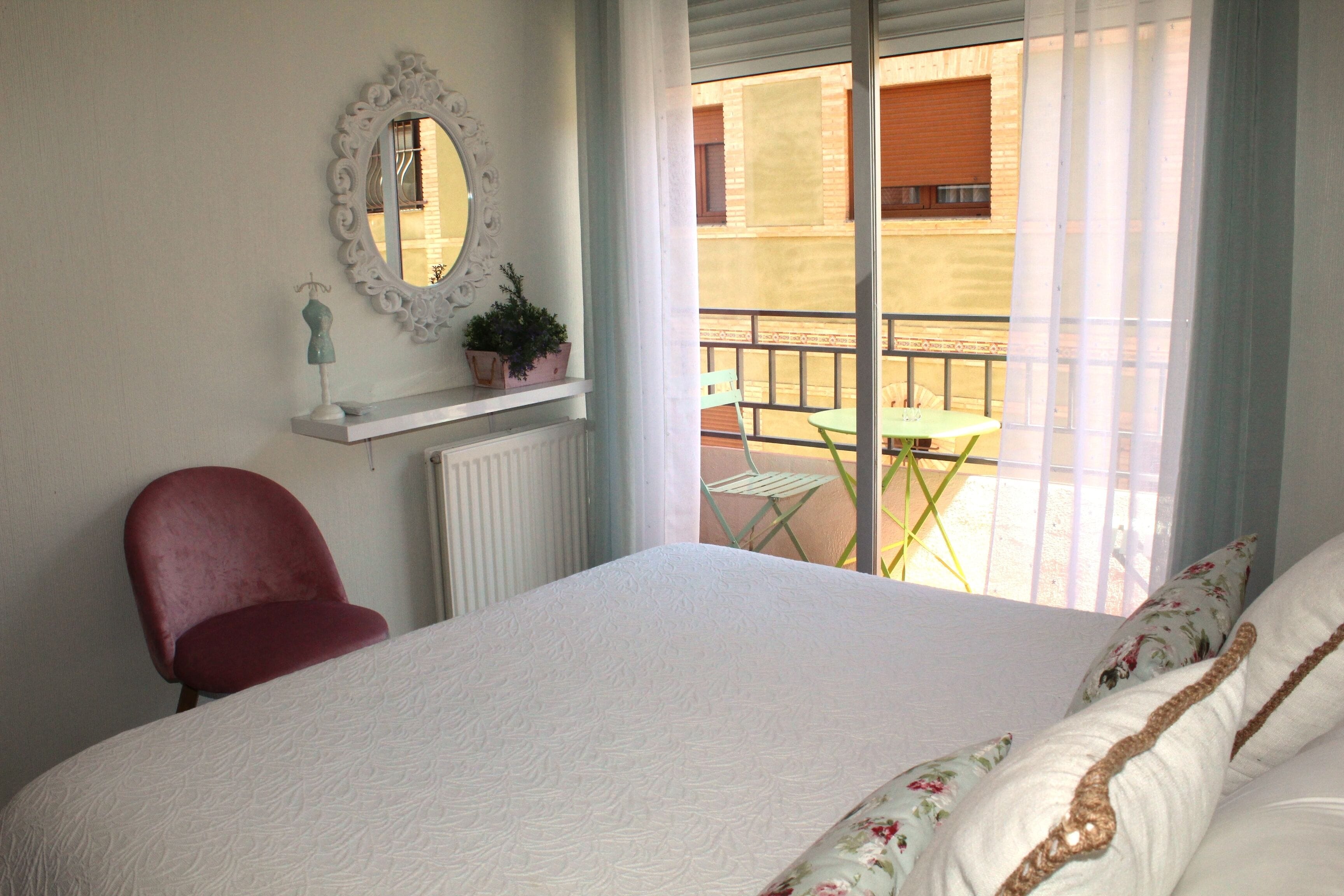 A bedroom with a large bed, a red chair, a small table, a mirror, and a balcony with a table and chairs.