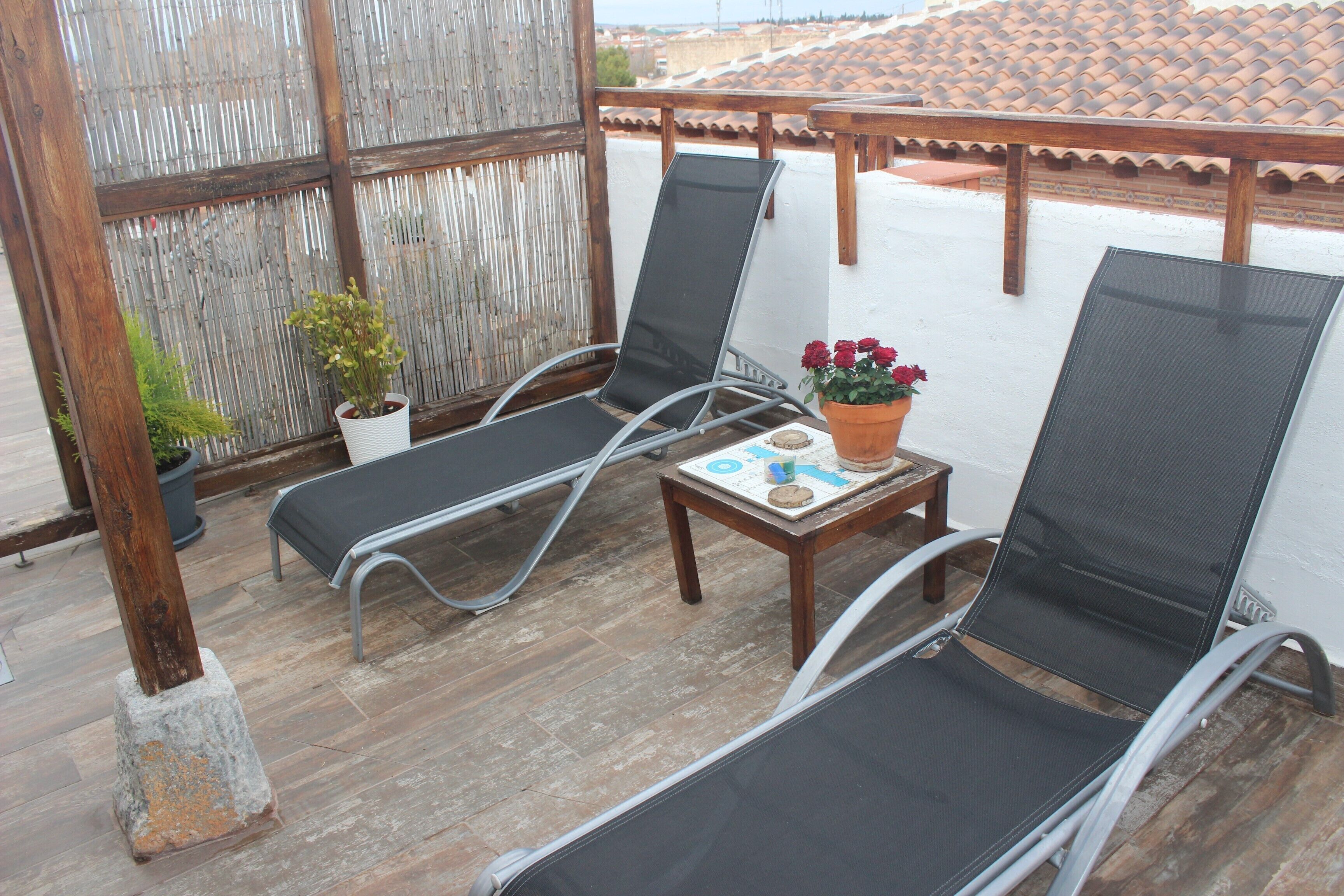 A balcony with two lounge chairs, a small table with a potted plant and a flower vase, and a wooden railing.