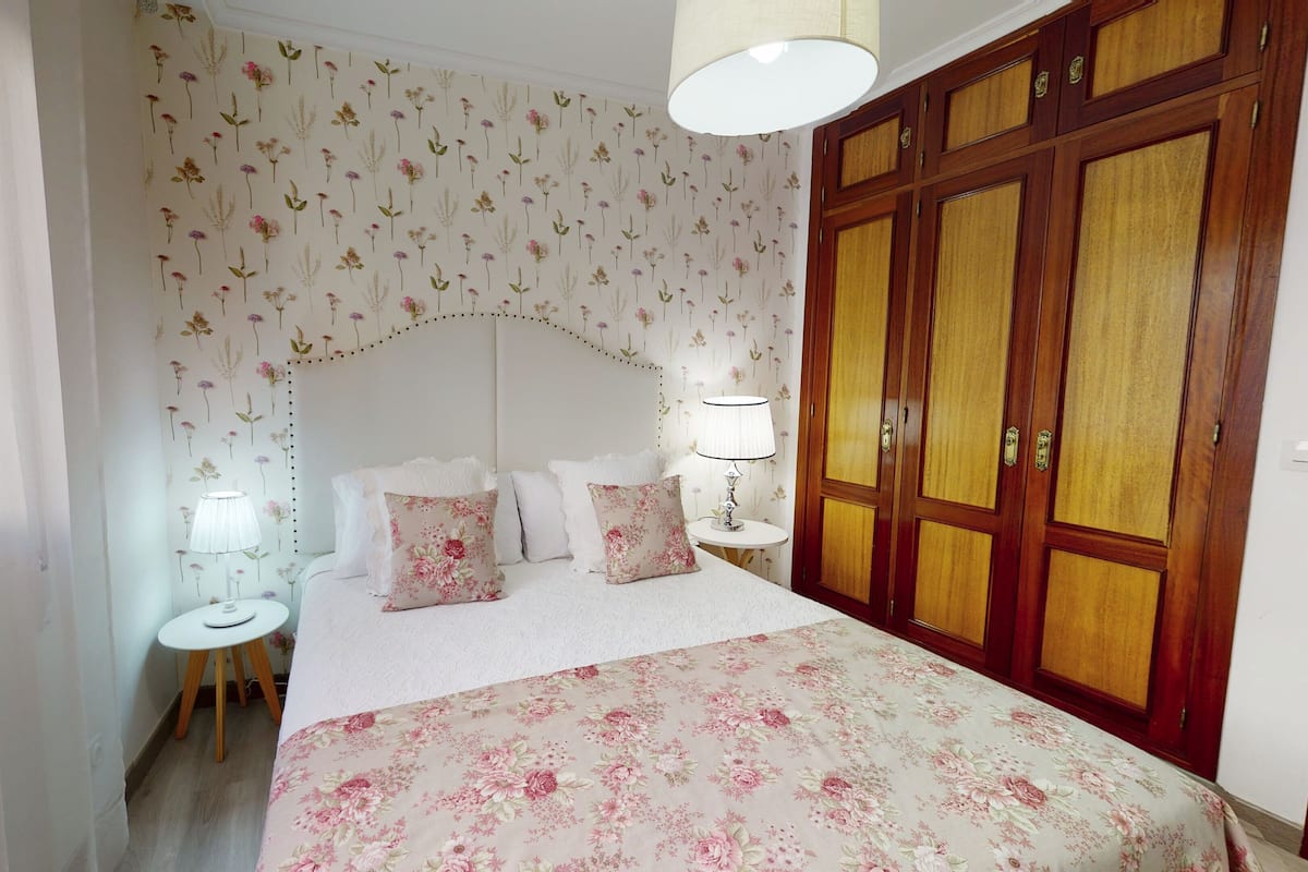 A bedroom with a floral wallpaper, a wooden wardrobe, a bed with floral bedding, two bedside tables with lamps, and a window with blinds.