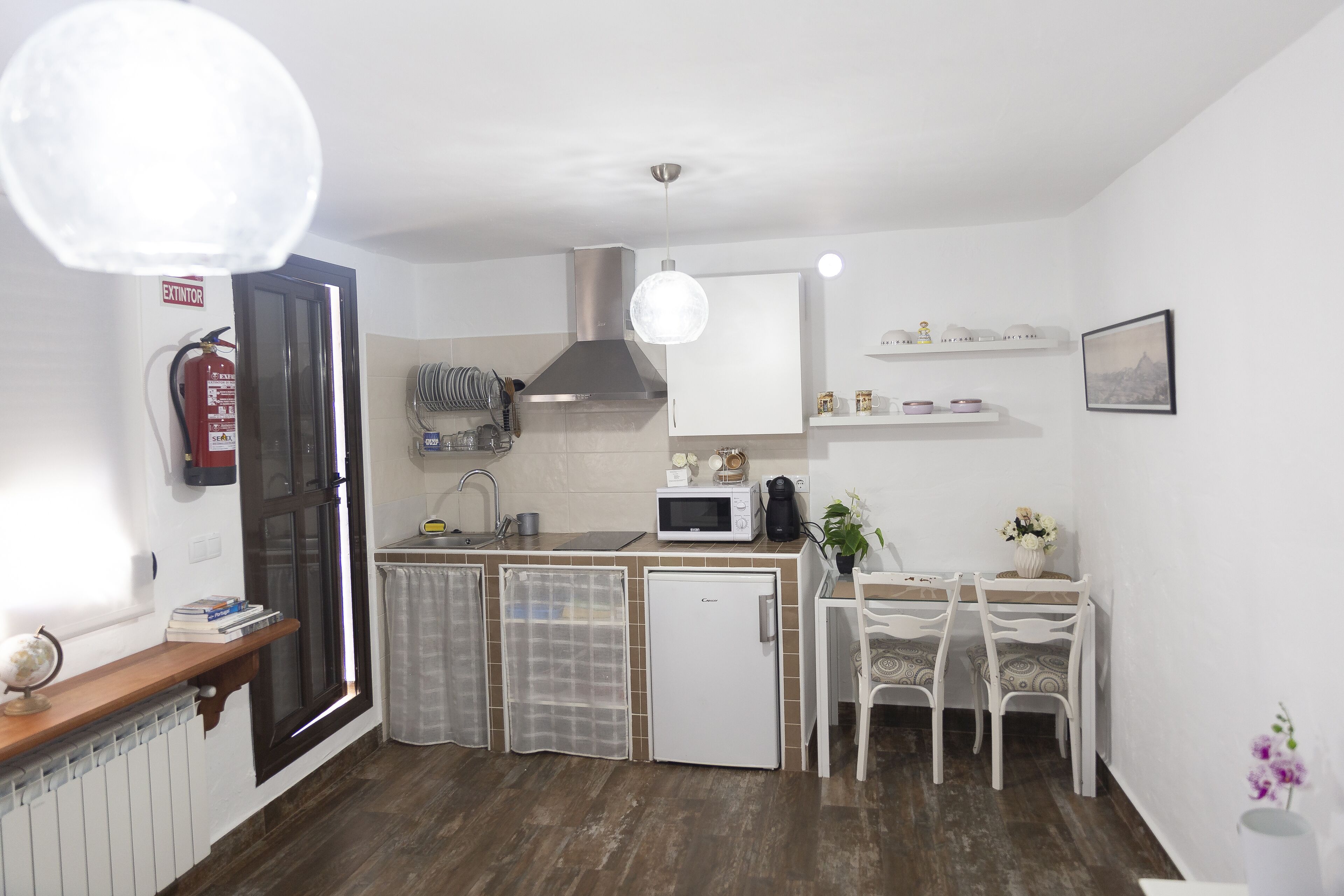 A compact kitchen with a refrigerator, microwave, and sink. There is a dining table with chairs and a small cabinet above it.
