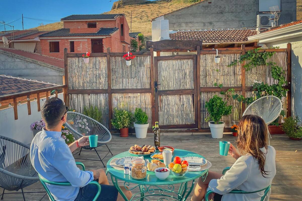A patio with a round table set for two, surrounded by metal chairs, with a dog and potted plants.