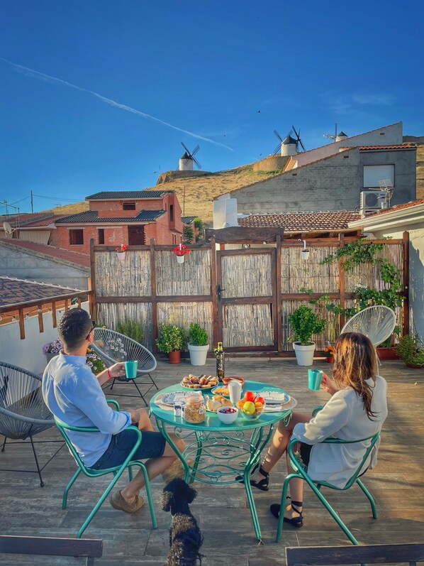 A patio with a round table set for two, surrounded by metal chairs, with a dog and potted plants. - Apartamentos OnceMolinos (Consuegra)