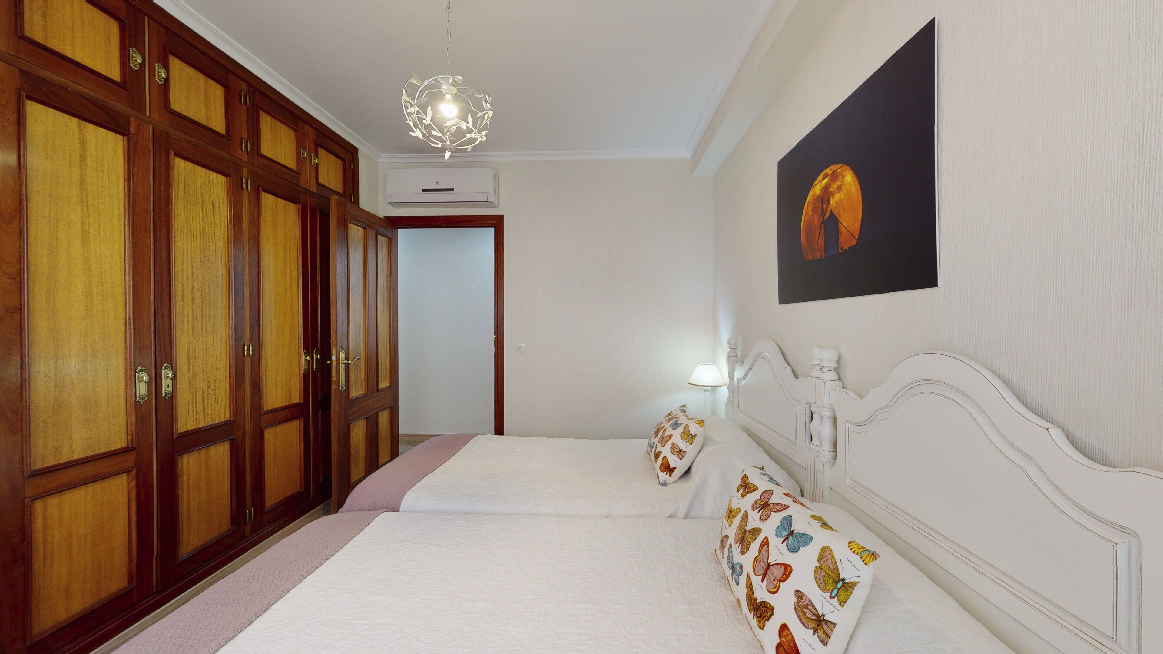 A hotel room with a large wardrobe, a bed with butterfly-patterned pillows, a wall-mounted painting, and a chandelier.