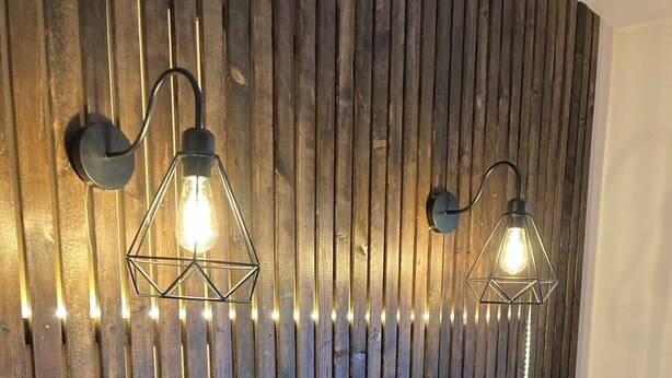 Une pièce avec un mur recouvert de lambris en bois, deux lampes fixées au mur et une couvre-lit à motifs.