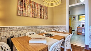 A dining area with a wooden table, white chairs, a woven light fixture, and a colorful abstract painting on the wall.