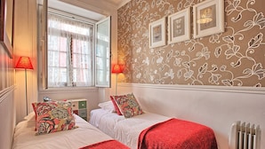 A room with two beds, red blankets, a radiator, and floral wallpaper.