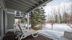 Eine überdachte Veranda mit zwei weißen Adirondack-Stühlen, einer Holzterrasse und einer verschneiten Landschaft mit Bäumen im Hintergrund.