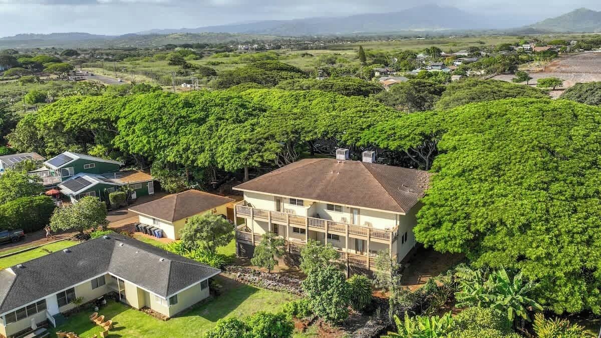 A large, two-story house with a balcony, surrounded by lush greenery and a hilly landscape.