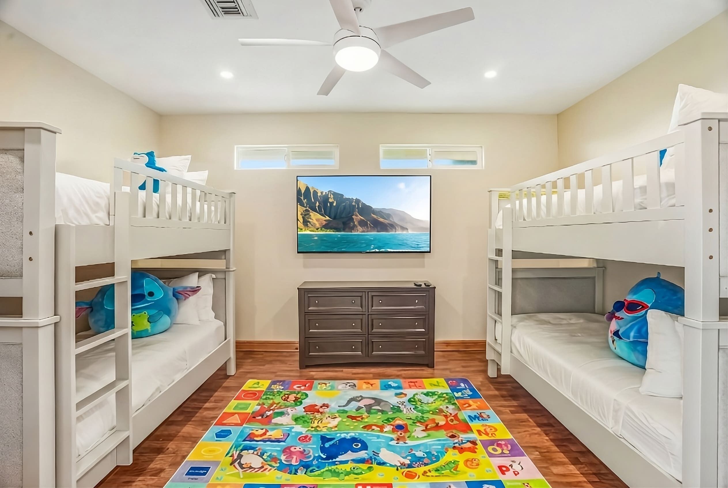 Une chambre d’enfant avec des lits superposés, un ventilateur de plafond, une commode et un tapis coloré représentant l’alphabet.