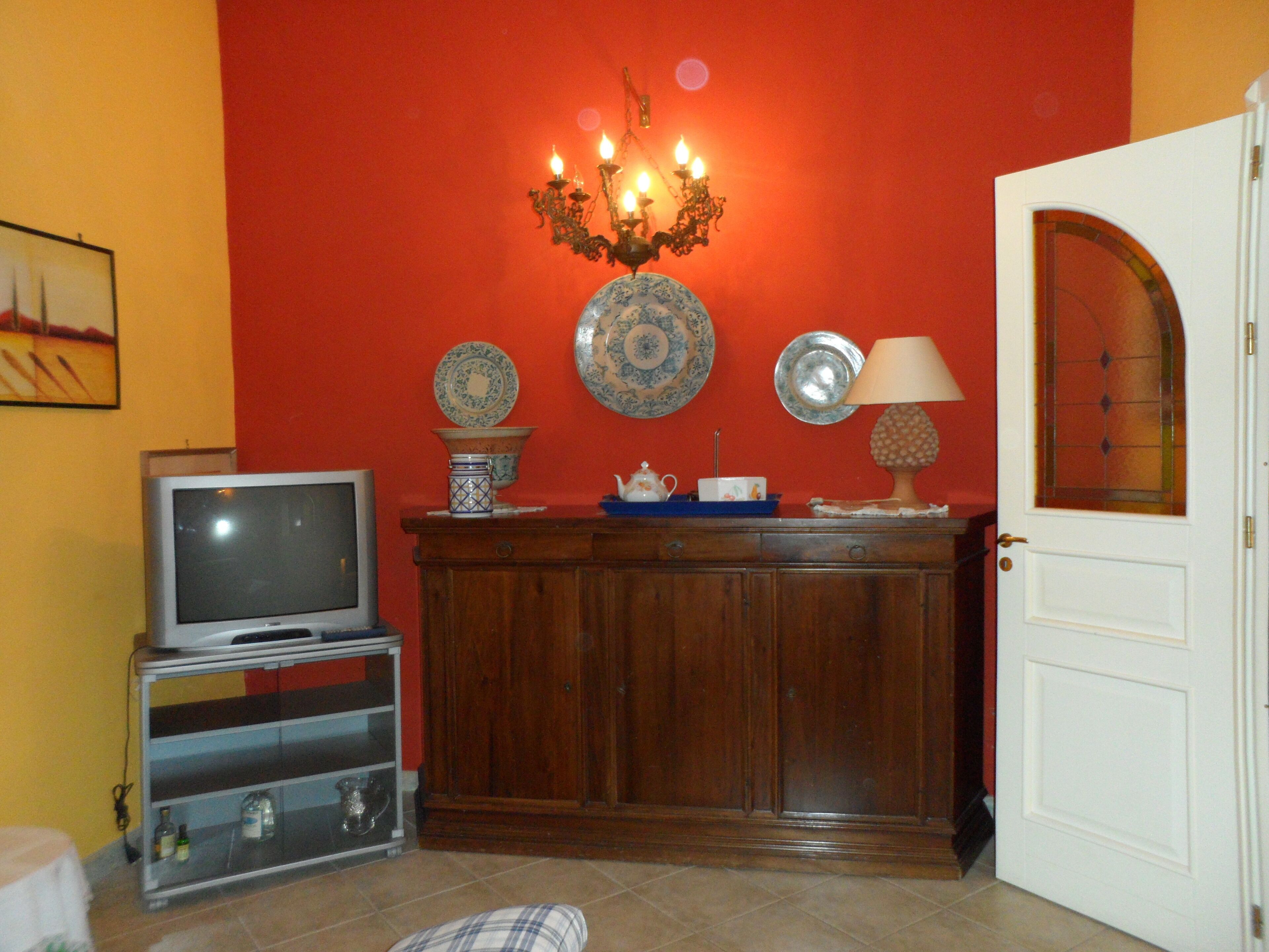 Una stanza con una televisione su un mobile, un armadio decorato con piatti ornamentali, un lampadario e una porta con un pannello di vetro.