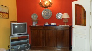 Een kamer met een televisie op een standaard, een kast met decoratieve borden, een kroonluchter en een deur met een ruit.