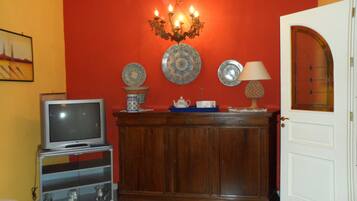 Una stanza con una televisione su un mobile, un armadio decorato con piatti ornamentali, un lampadario e una porta con un pannello di vetro.