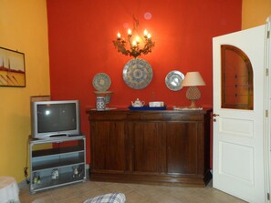 A room with a television on a stand, a cabinet with decorative plates, a chandelier, and a door with a glass panel.
