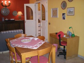 Un comedor con una mesa dispuesta para cuatro, una silla roja, un escritorio de madera con una silla, y una puerta que da a la cocina.