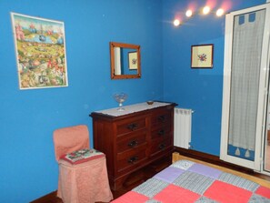 Un dormitorio con pared azul, cómoda de madera, silla rosa, cama con colcha, radiador y dos cuadros enmarcados.