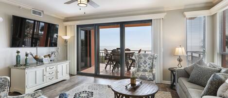 A spacious living room with a large sliding glass door leading to a balcony with a view of the ocean. There is a ceiling fan, a flat-screen TV mounted on the wall, and a white cabinet with decorative items.