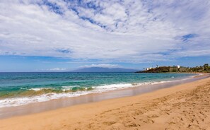 A beach with clear turquoise waters, a sandy shore, and a distant mountain range under a partly cloudy sky.