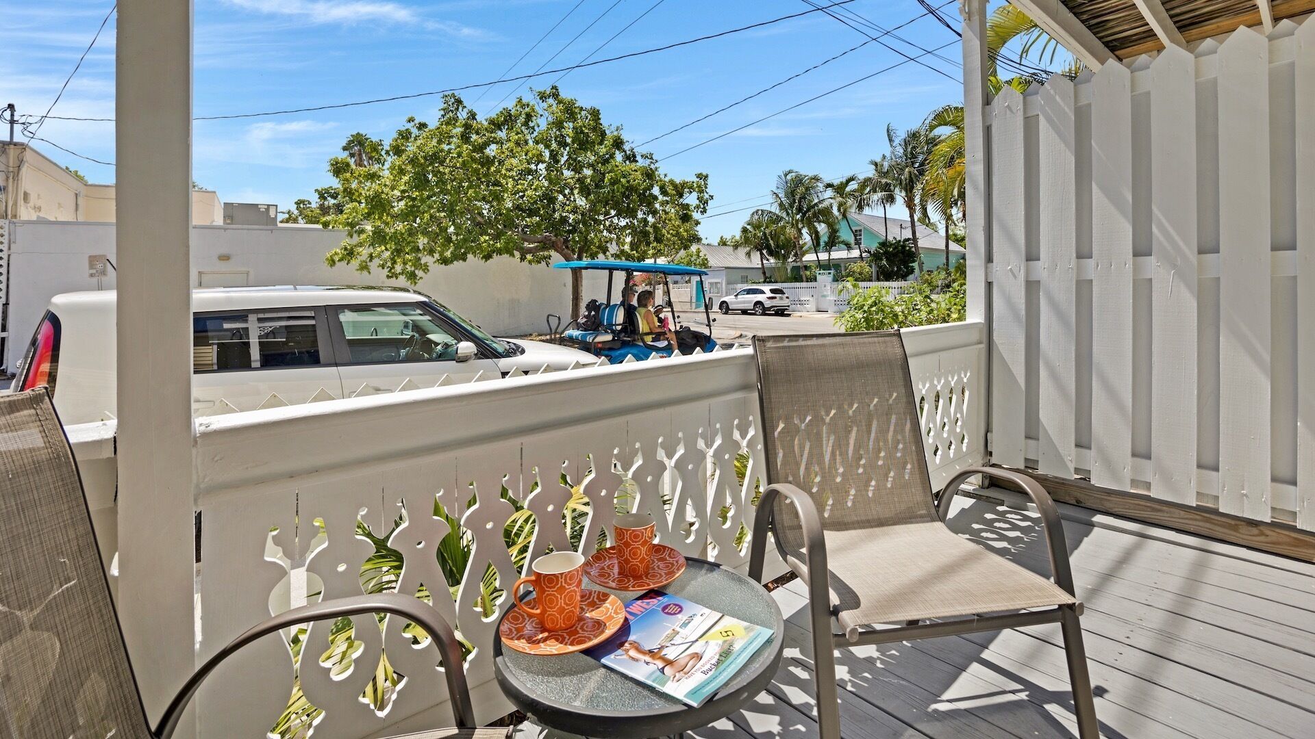 Un espacio al aire libre cubierto con dos sillas y una mesa, una cerca con detalles decorativos y vista a una calle con coches aparcados y un carrito de golf.
