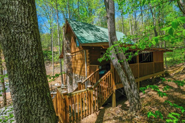 A wooden cabin with a green roof, surrounded by trees, featuring a wooden staircase leading to a deck. - 1BR log cabin with pool table, hot tub, and convenient location! (Sevierville)