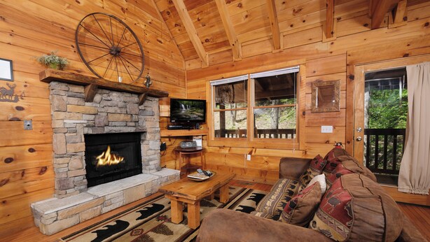 A cozy cabin interior with a stone fireplace, wooden walls, a large window, and a comfortable seating area. - POOL TABLE, HOT TUB, AND JACUZZI TUB in a CONVENIENT location! (Sevierville)