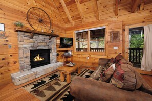 Cozy cabin features a stone fireplace, comfortable sofa, and TV on a wooden stand, with a window offering a forest view.