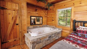 A wooden cabin bedroom with a stone bathtub, a bed with a patterned blanket, and a window with a forest view. - POOL TABLE, HOT TUB, AND JACUZZI TUB in a CONVENIENT location! (Sevierville)