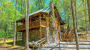 A cabin with a stone foundation, wooden upper structure, and a metal roof, surrounded by a forest. - POOL TABLE, HOT TUB, AND JACUZZI TUB in a CONVENIENT location! (Sevierville)