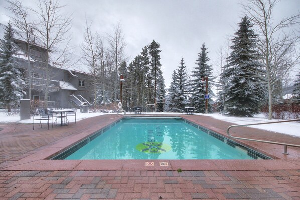 A rectangular pool with a brick border, surrounded by snow-covered trees and a building with a balcony.