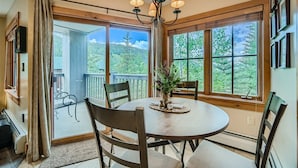 A dining area with a round table, chairs, a chandelier, and large windows with a view of greenery. - Bright Corner Unit in River Run/Walk to the Slopes/Pool and Hot Tub Access (Keystone)