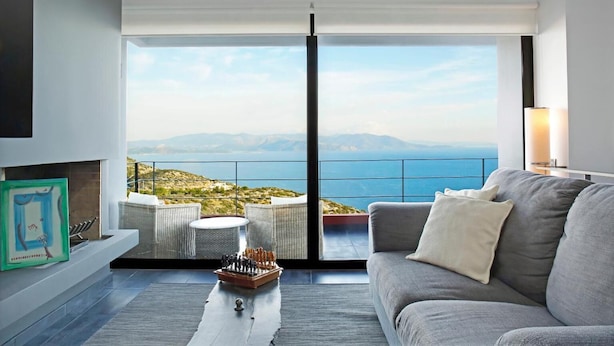 A modern living room with a grey sofa, a small round table, and a chessboard. A large window offers a view of the sea and distant mountains. - Sea View Villa Schinias (Marathon)