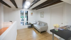 Un salon moderne avec un plafond en bois, un canapé gris, une petite table sur laquelle est posé un vase de fleurs et une fenêtre avec des stores.