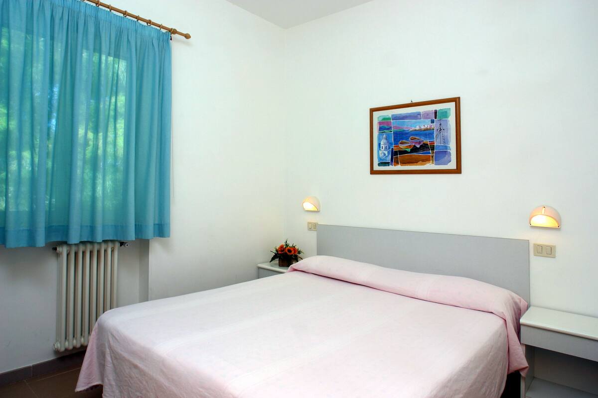 Un dormitorio con una cama, una mesita de noche, un cuadro en la pared y una ventana con cortina azul.
