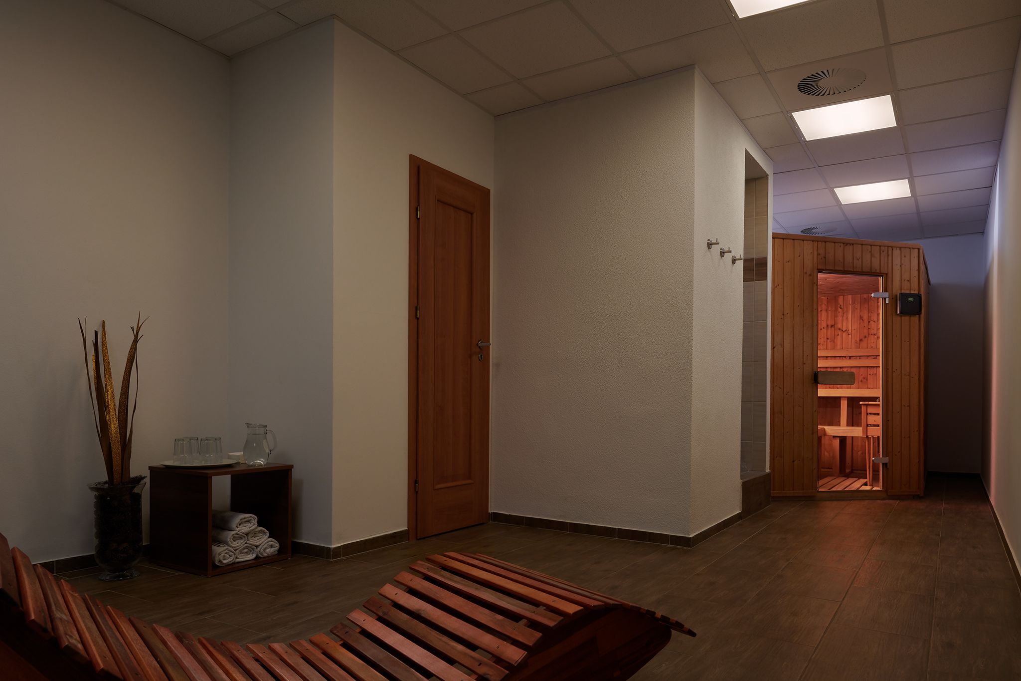 Ein Zimmer mit einer hölzernen Bank, einem kleinen Tisch mit Vase und Gläsern sowie einer Tür zum Saunabereich.