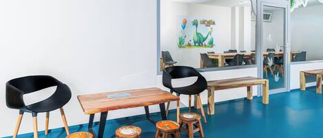 Una habitación con piso azul, mesas de madera, sillas negras y de madera, y un mural con una fiesta de dinosaurios.