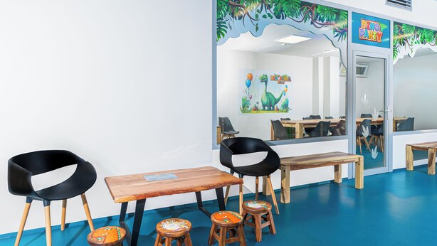 Una habitación con piso azul, mesas de madera, sillas negras y de madera, y un mural con una fiesta de dinosaurios.
