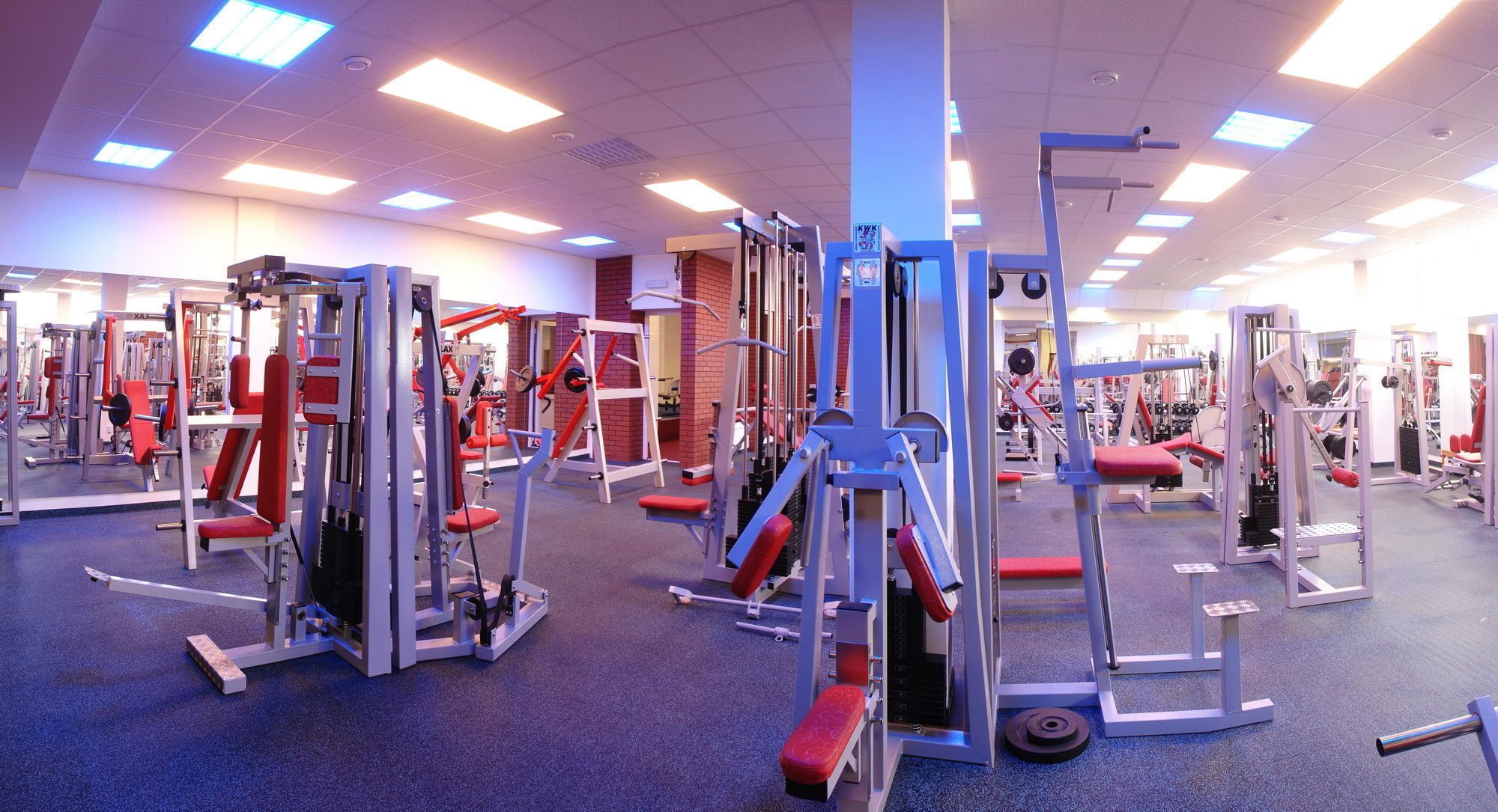 Un gimnasio con diferentes máquinas de ejercicio, un pilar azul y un banco rojo.