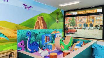 Ein farbenfroher Spielbereich mit Dinosaurier- und Tierdekorationen, einem Klavier und einem Fenster mit Blick auf eine Stadtstraße.