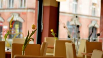 Interior de restaurante con mesas y sillas, un jarrón con una flor solitaria, y vista a un edificio de fachada rosa y ventanas.