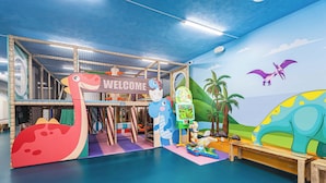 Een binnenspeelruimte met een 'Welcome' bord, een speelconstructie met dinosaurus thema en een muurschildering met een vliegende vleermuis.