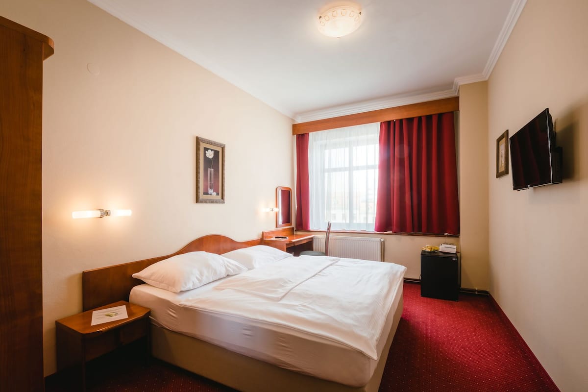 Habitación de hotel con cama, mesita de noche, lámpara, ventana con cortinas y televisión en la pared.