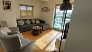Een woonkamer met een grijze bank, een grijze fauteuil, een salontafel en een groot raam met uitzicht op een stadsbeeld.