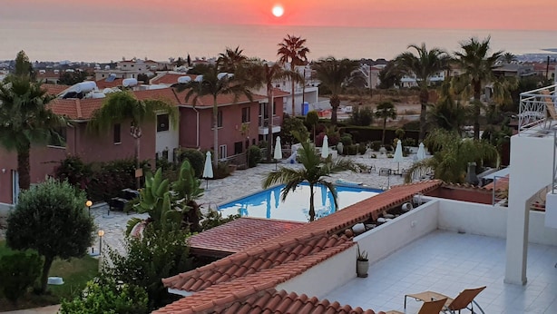 Ein Poolbereich auf dem Dach mit Blick auf den Sonnenuntergang über dem Meer, umgeben von Palmen und Gebäuden mit roten Ziegeldächern.