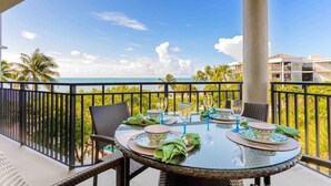 A balcony with a glass table set for dining, overlooking a beach with palm trees and a clear sky.