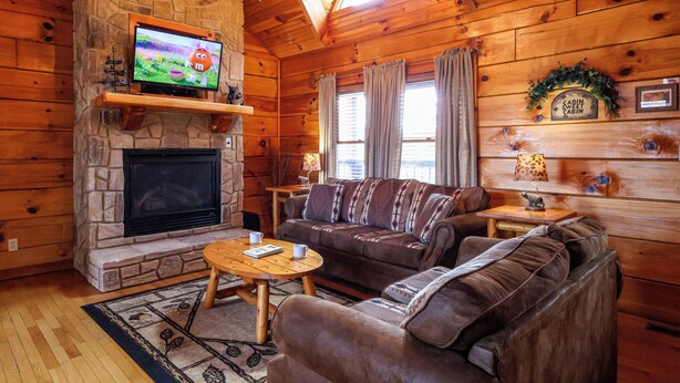 A cozy cabin interior with a stone fireplace, a television mounted above it, a brown sofa, and a small table with a lamp. - One of the coziest log cabins in the Smokies! With a hot tub and a jacuzzi tub! (Pigeon Forge)