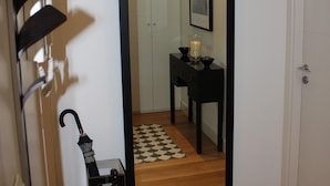 Un pasillo con piso de madera, un espejo de marco negro que refleja una habitación con piso a cuadros, una mesita con una vela y un paraguas negro apoyado contra la pared.