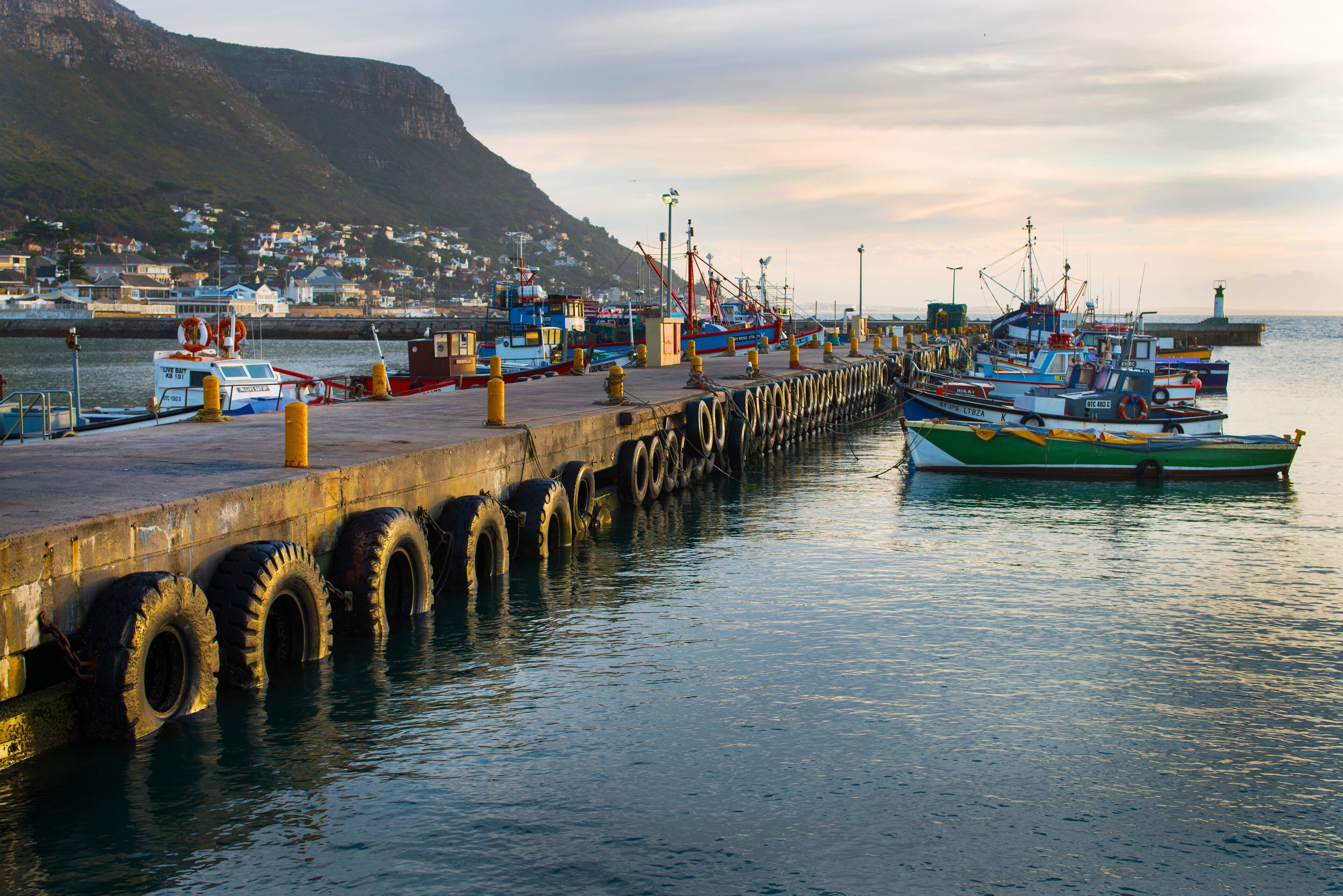 A pier with boats docked, a lighthouse in the background, and a town across the water.